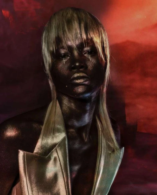 Striking beauty portrait of a dark-skinned woman with platinum blonde hair, metallic gold makeup, and a gold jacket, set against a red background.