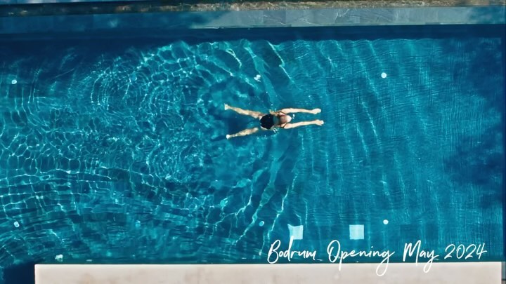 Overhead view of a woman swimming in a pool with turquoise water, creating ripples, and a sign indicating Bodrum's opening in May 2024.
