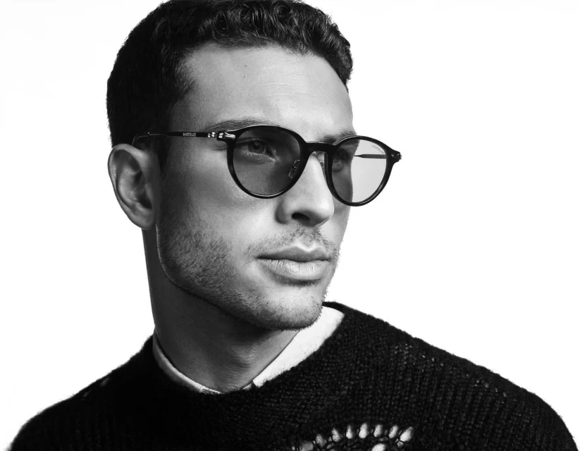 Black-and-white portrait of an adult male wearing round sunglasses and a black sweater against a bright white background.