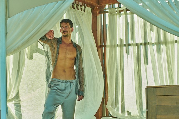 Attractive shirtless man wearing a sheer shirt and silk pants in a sheer curtained cabana looking away from the camera.