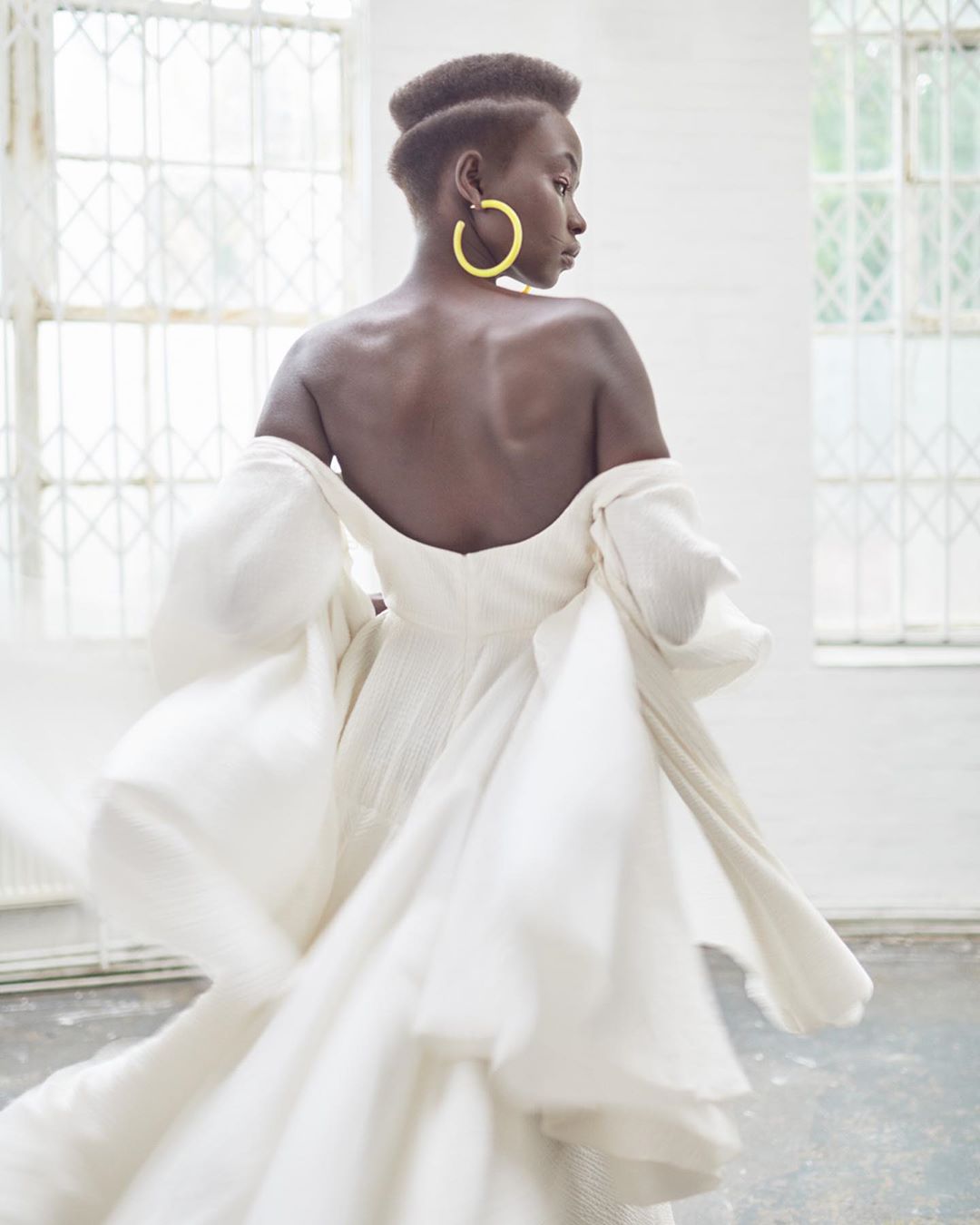 Woman in flowing white off-the-shoulder dress with oversized sleeves, golden hoop earrings, and short dark hair turning away from the camera.