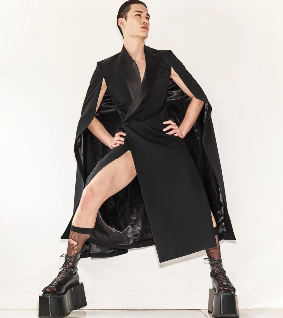 A man poses in a black wrap coat and platform boots against a white background, showcasing a high-fashion editorial look.