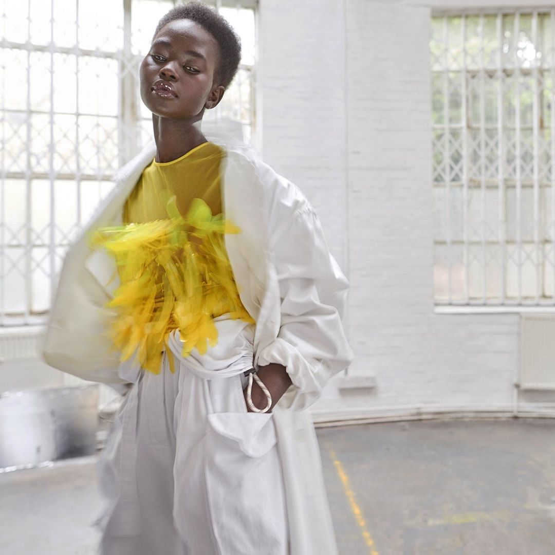 Fashion shot of Black female model in white pantsuit and yellow floral top posing in a bright white industrial setting with large windows.