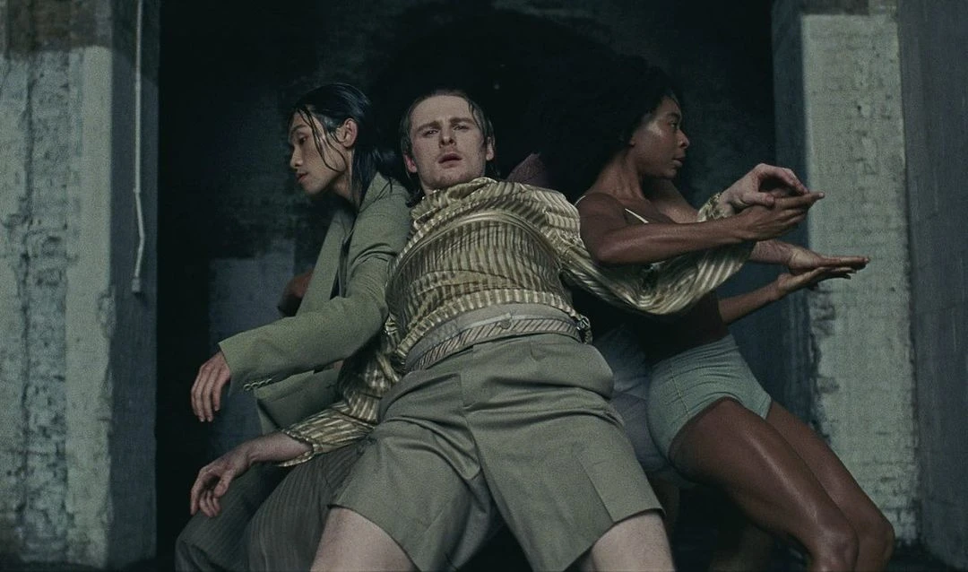Three people in a dimly lit, concrete-walled space. A man in striped shirt is centered between two other figures in semi-formal attire.