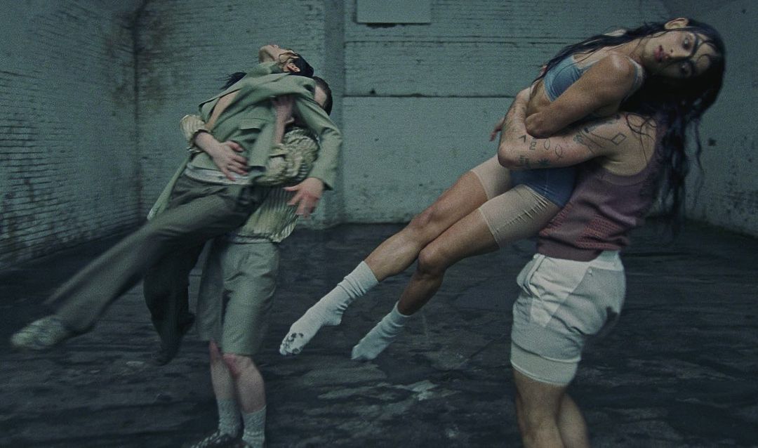 Two pairs of dancers lifting each other in a spacious industrial warehouse with pale brick walls and a concrete floor