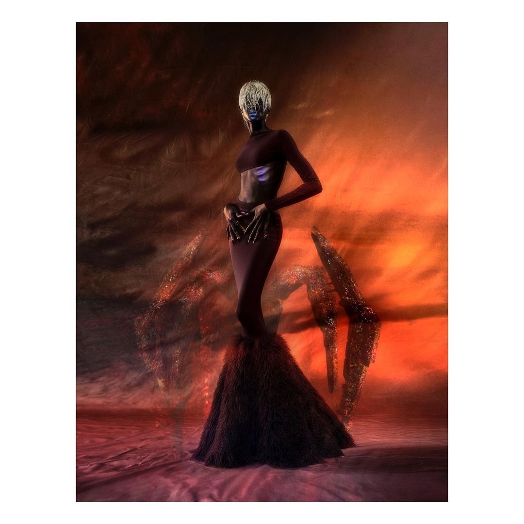 Elegant woman in a maroon gown with a dramatic feathered skirt poses against a swirling orange and black abstract background in a studio setting.