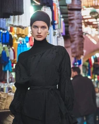 Model in black turtleneck and headband with gold earrings wearing dark top against market background