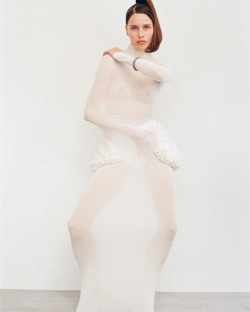 A slender woman with a ponytail poses in a full-body sheer white bodysuit and intricately designed white accent pieces against a white wall.