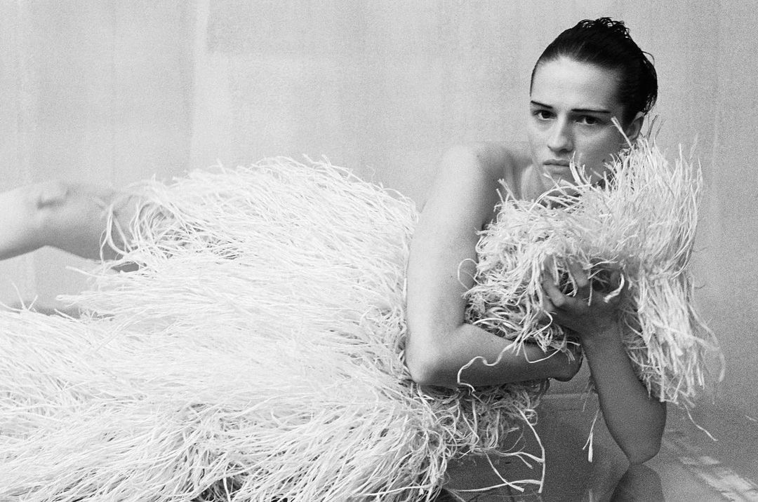 Monochrome portrait of an adult woman partially covered in shredded paper, looking at camera in a minimalist indoor setting.
