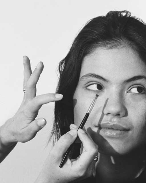 Woman receives makeup application from a makeup artist using a brush, set against a clean, minimalist white backdrop in a beauty setting.