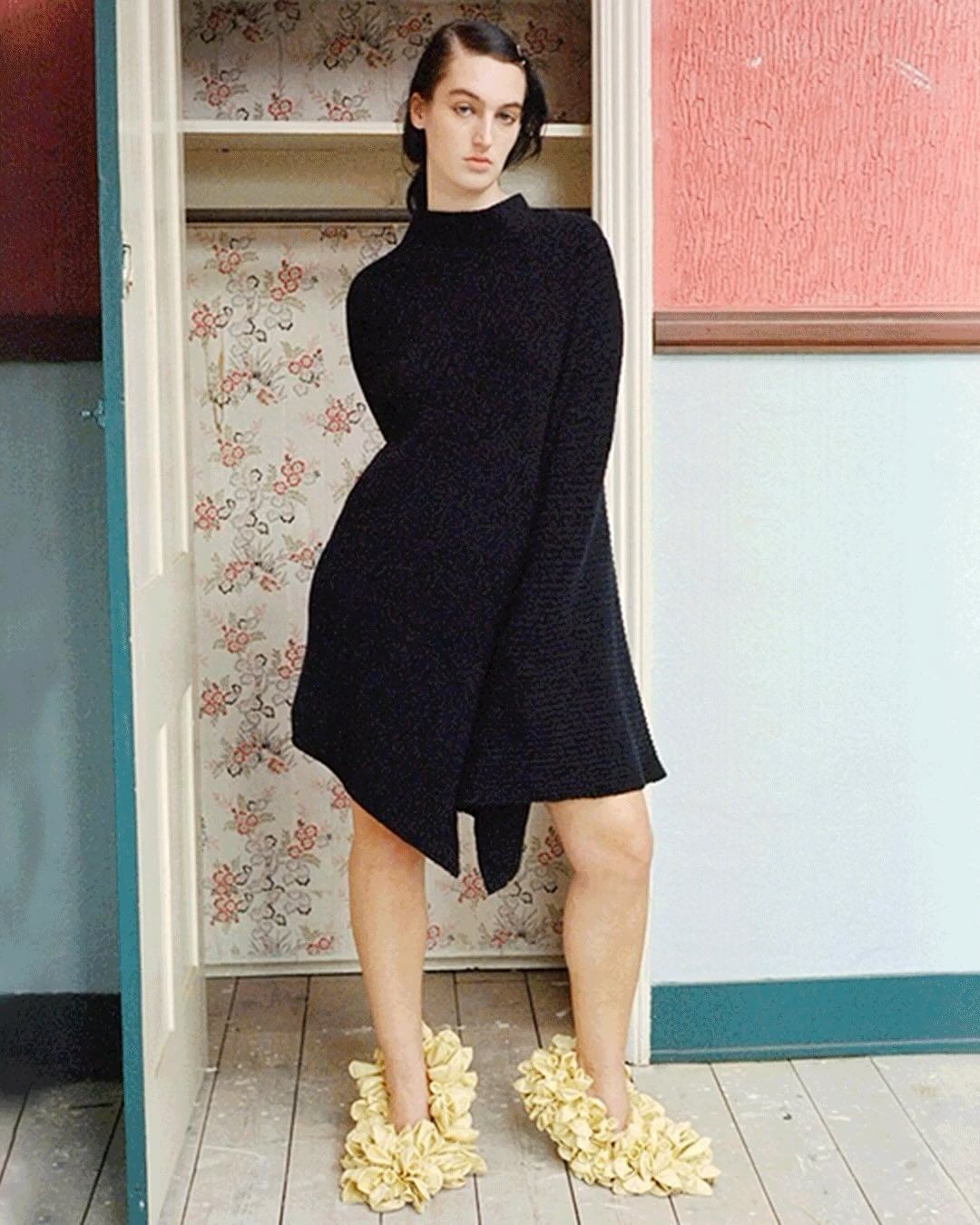 Person in a black dress stands in a doorway wearing unique beige floral shoes, posing in front of a floral closet and pink wall.