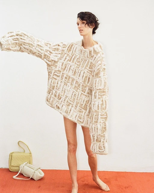 An adult female model poses indoors wearing a large beige woven sweater, standing on a red carpet with a handbag and purse.