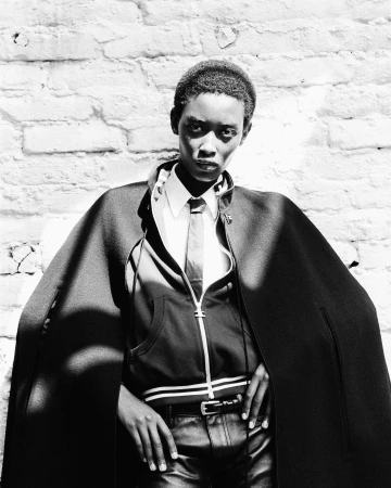 Black and white portrait of a model in a dark cape, zip-up jacket, and tie against a textured brick wall.