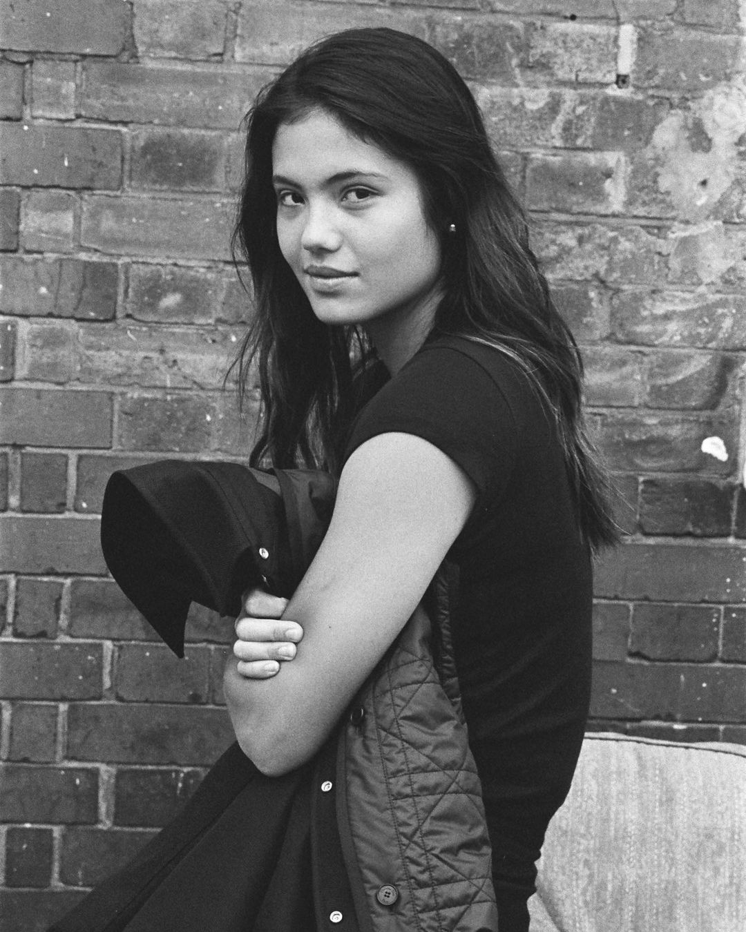 Black and white portrait of a woman with long dark hair, against a brick wall, holding a jacket, looking at the camera.