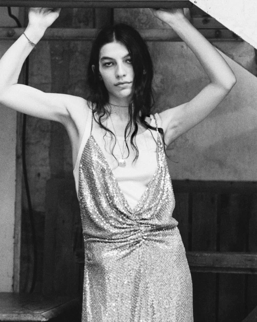 Monochrome portrait of a slender adult woman wearing a spaghetti strap dress with metallic fabric, indoors with dark background.
