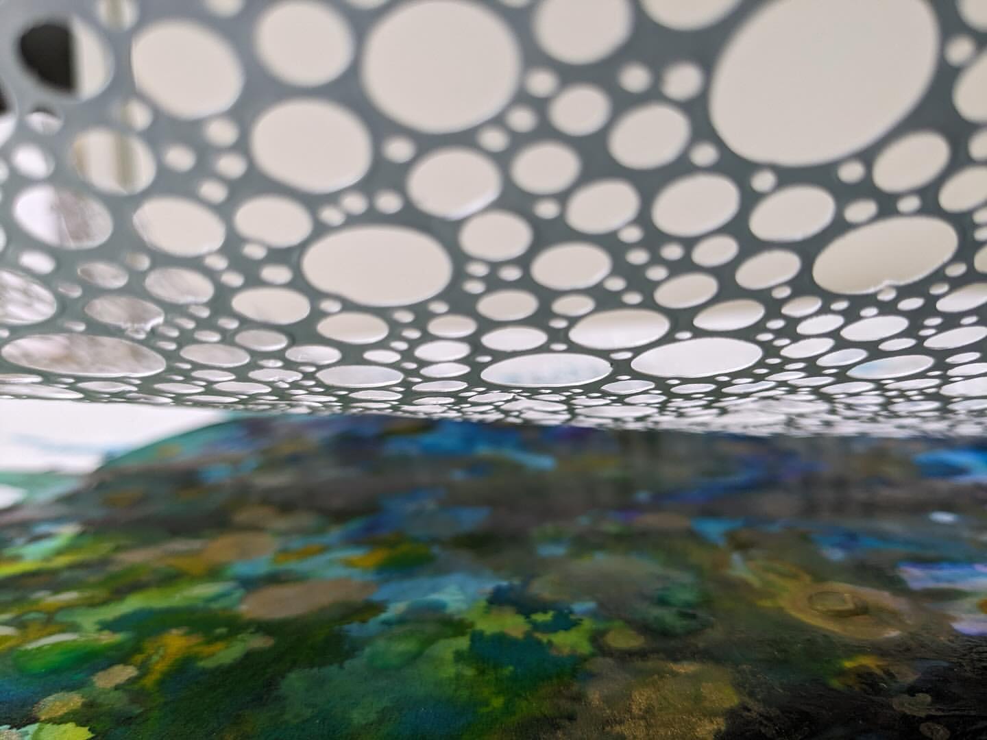 Abstract macro shot of a gray metal surface with many circular perforations, above a blurry colorful abstract painting.  