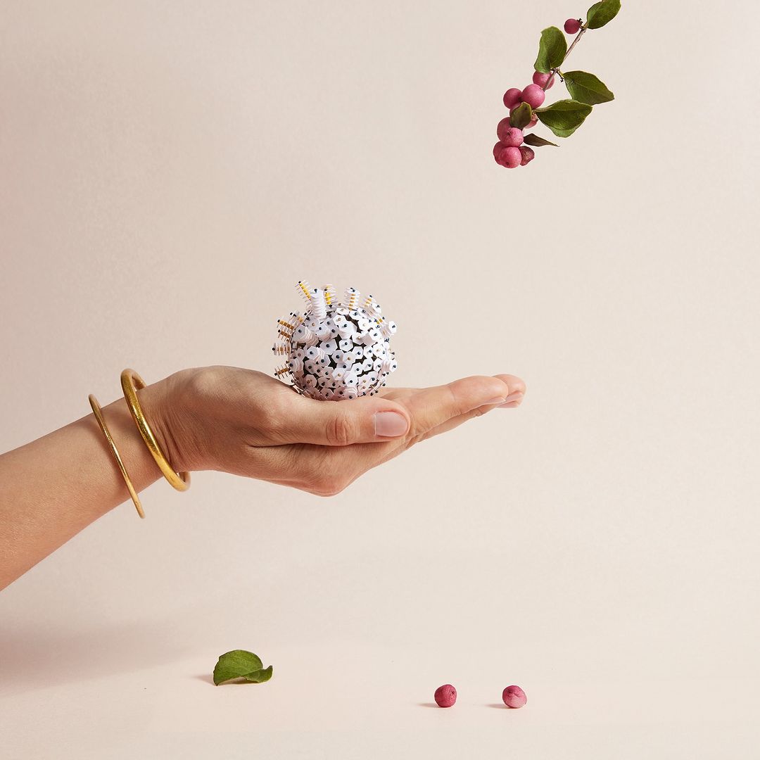 A woman's hand holds a decorative ball with white and black spots against a light background, accessorized with two gold bracelets.