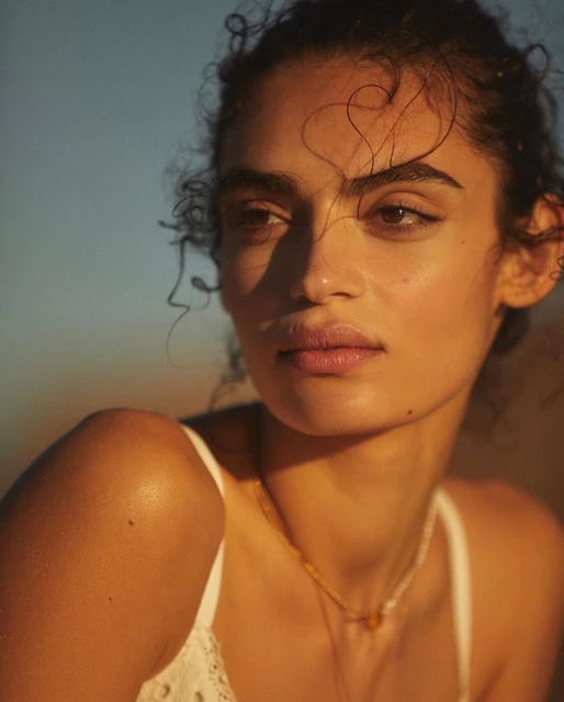 Portrait of a woman with curly hair and a white top, illuminated by warm light against a gradient blue and tan background.