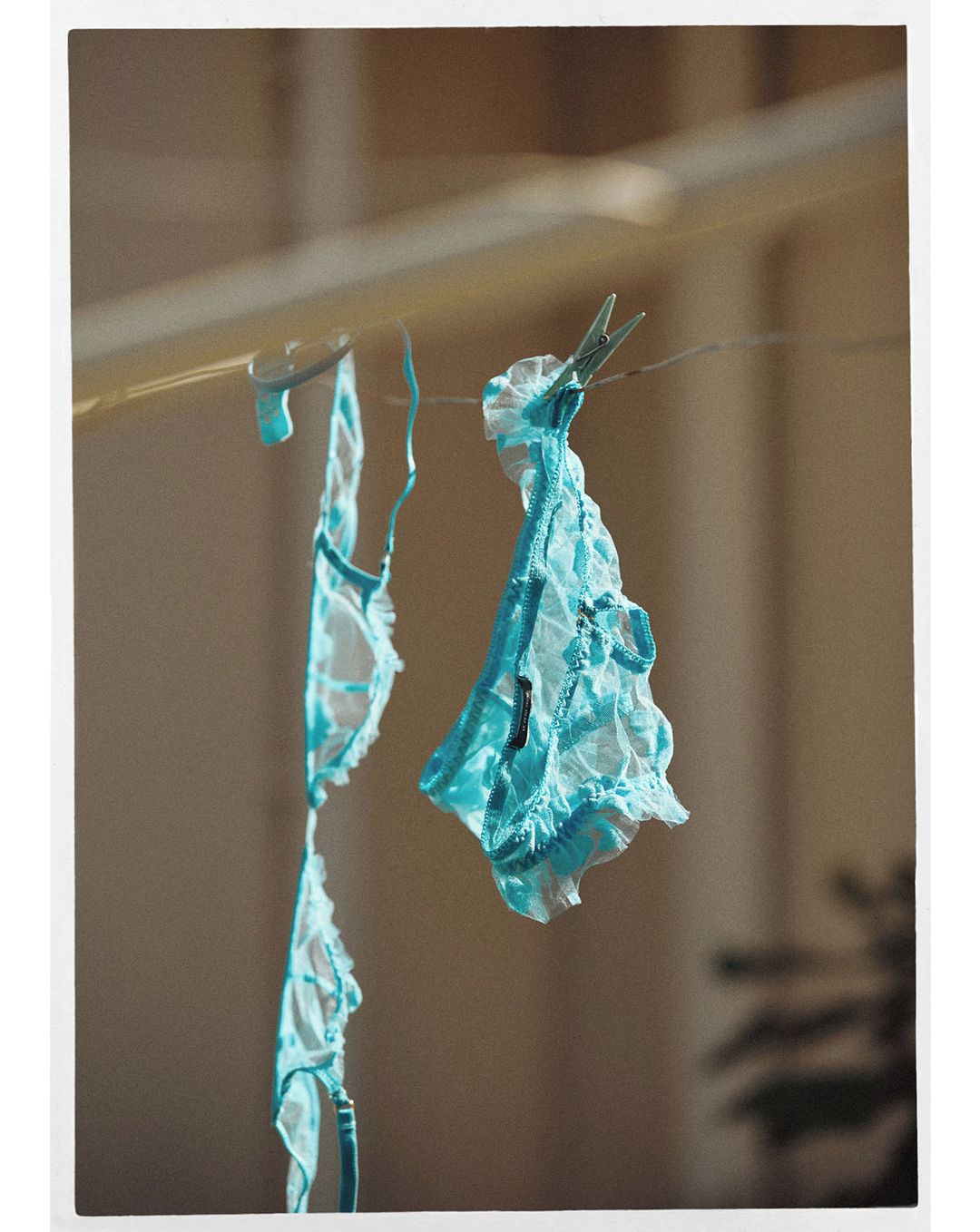 Delicate light blue lace lingerie hangs by clothespins from a wire in a sunlit, neutral indoor setting. Feminine fashion.