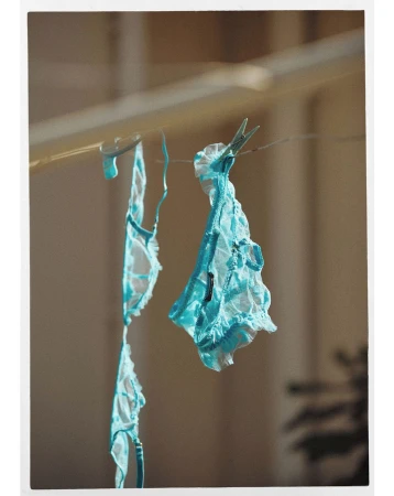 Delicate light blue lace lingerie hangs by clothespins from a wire in a sunlit, neutral indoor setting. Feminine fashion.