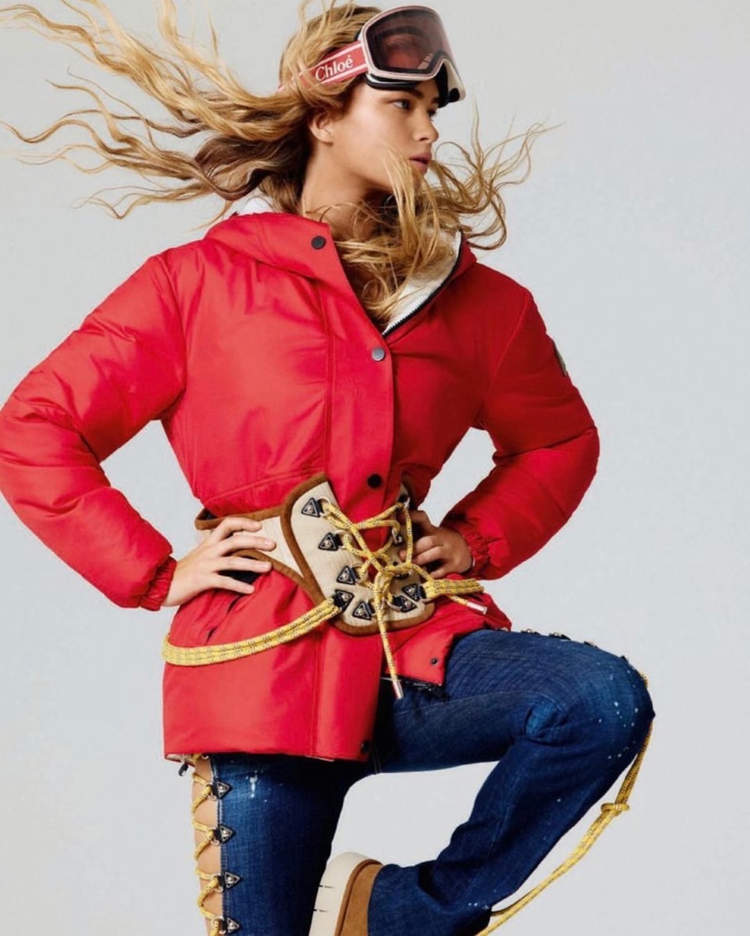 A woman poses in a red puffer jacket, ski goggles, and jeans with tan laces, creating a winter fashion editorial shot on a white background.