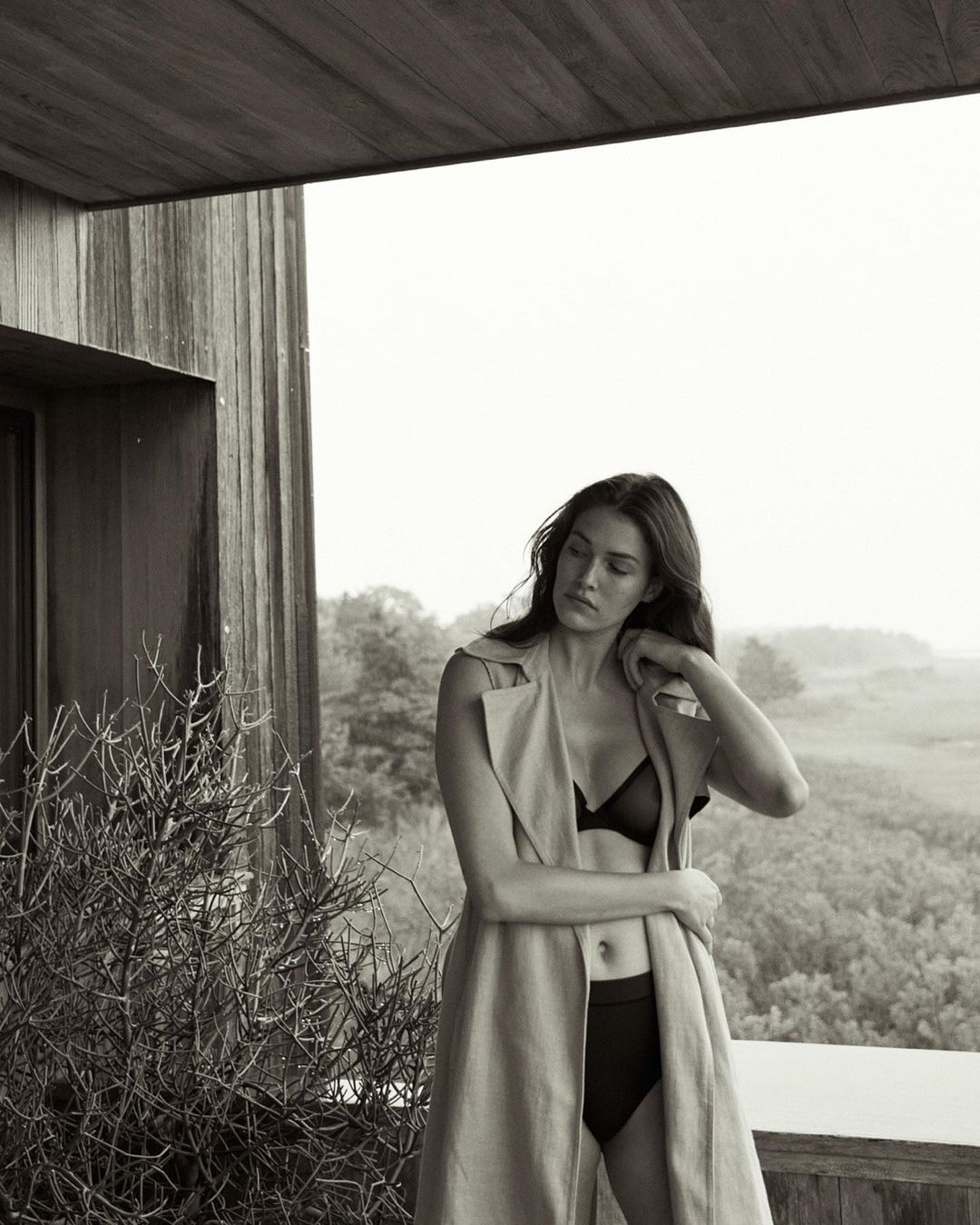 A woman in lingerie and a coat poses on a patio with foliage and greenery in the background in a black and white medium shot.