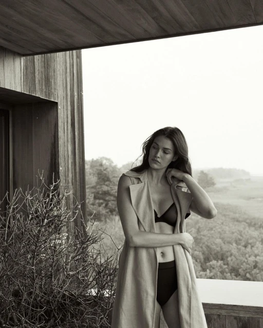 A woman in lingerie and a coat poses on a patio with foliage and greenery in the background in a black and white medium shot.