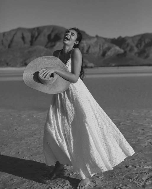 Woman in white dress laughing on beach with sun hat. Sunny day, monochrome shot of woman in dress with mountains in background.