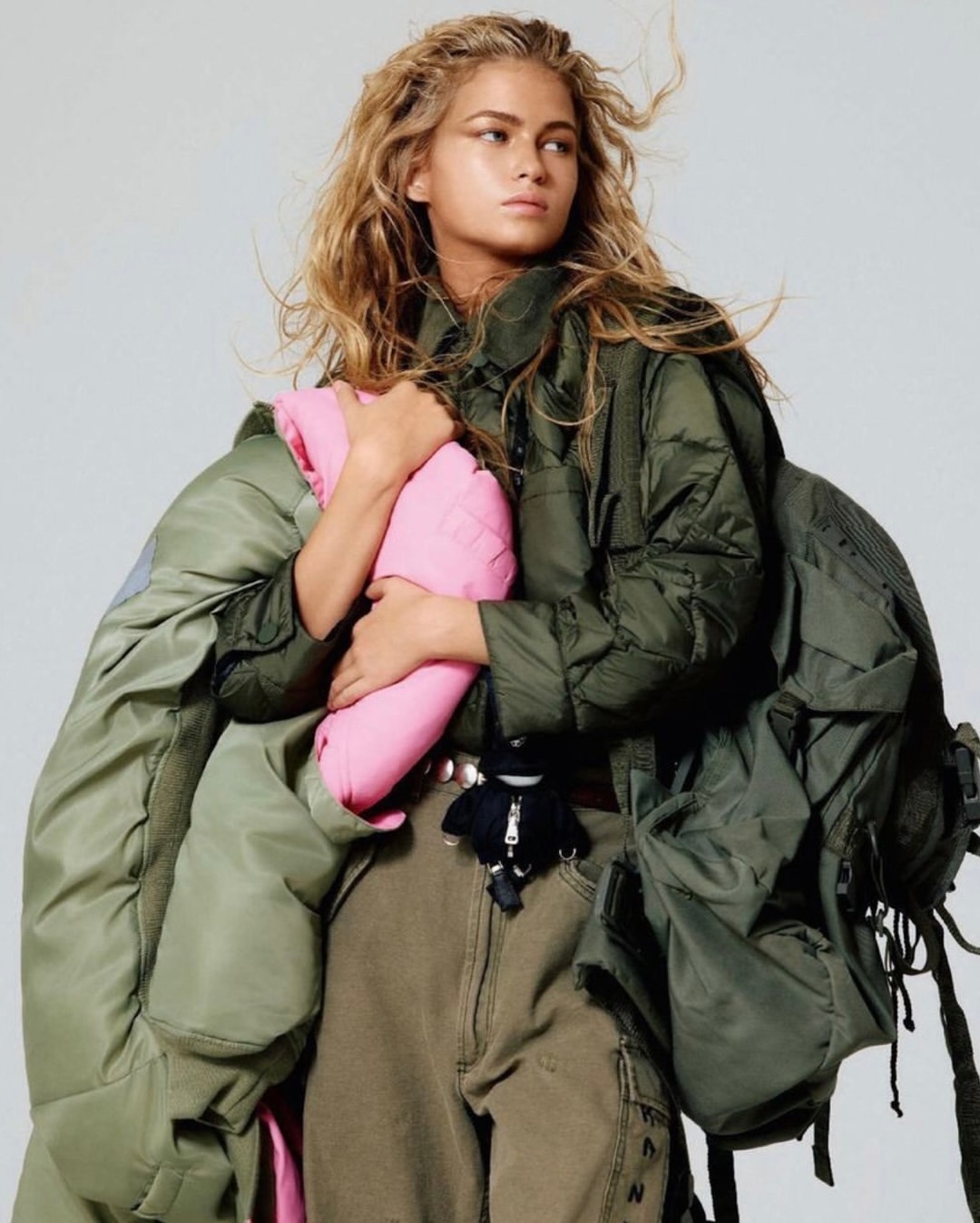 Fashion shot of a woman with blond hair holding a pink item, wearing cargo pants and a green jacket, standing indoors.