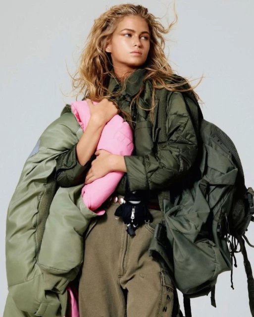 Fashion shot of a woman with blond hair holding a pink item, wearing cargo pants and a green jacket, standing indoors.