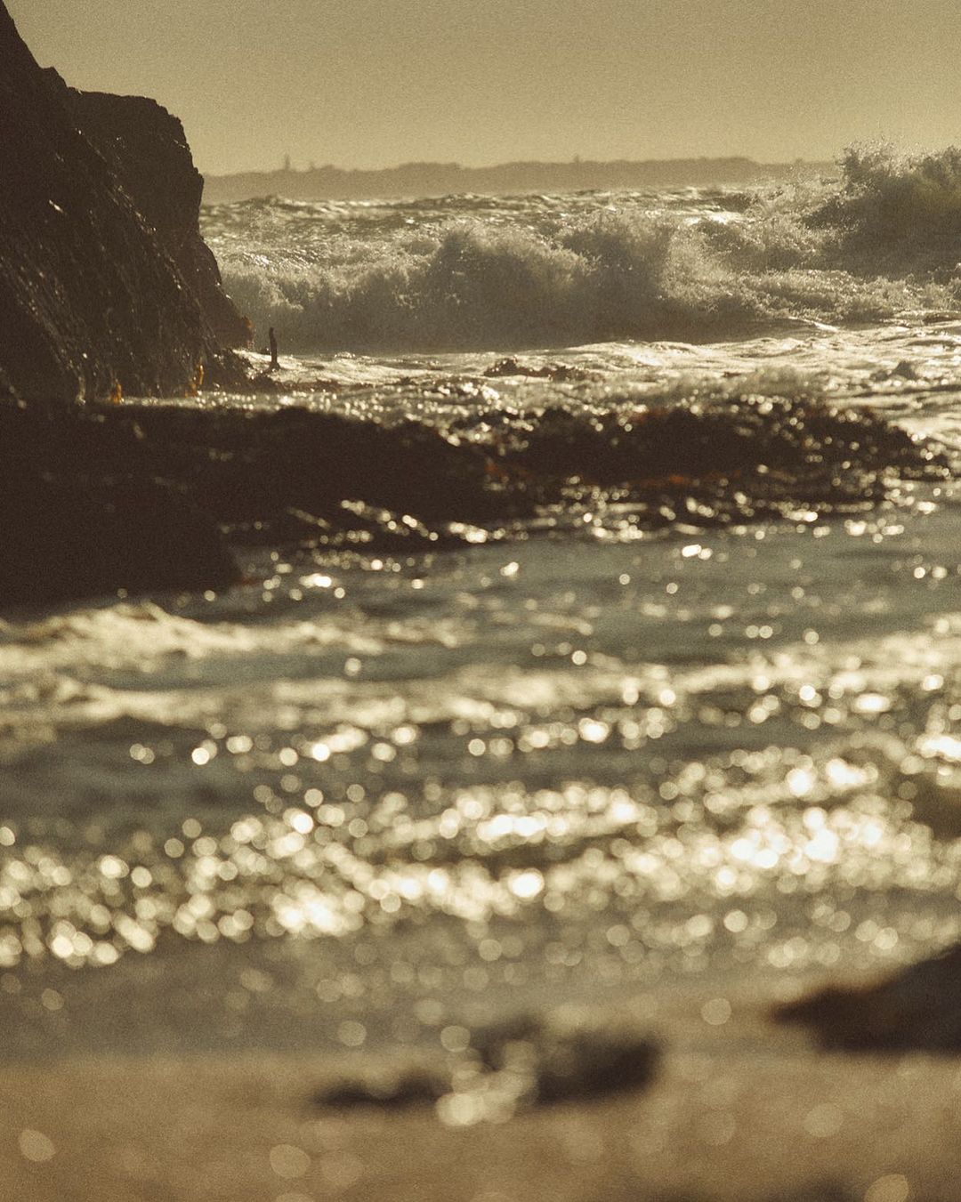 Ocean waves crash against a rocky coastline, with golden light reflecting off the water and illuminating the rugged landscape.