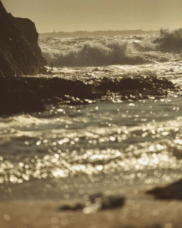 Ocean waves crash against a rocky coastline, with golden light reflecting off the water and illuminating the rugged landscape.