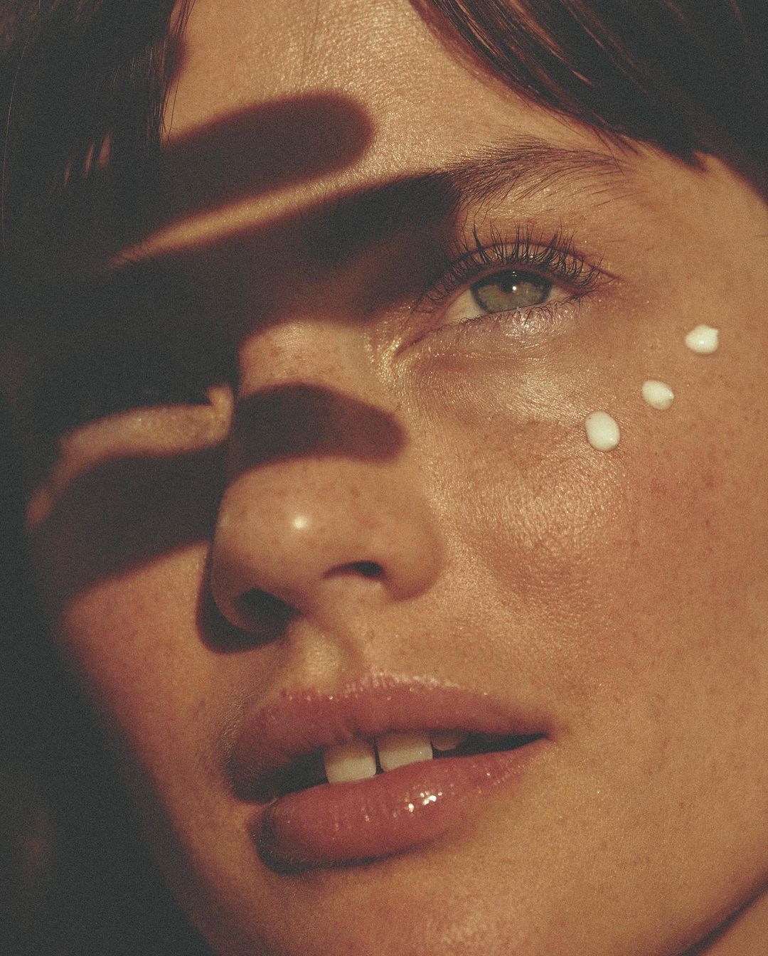Close-up portrait of a fair-skinned woman with cream dots under her eye, partially shaded face, and glossy lips.