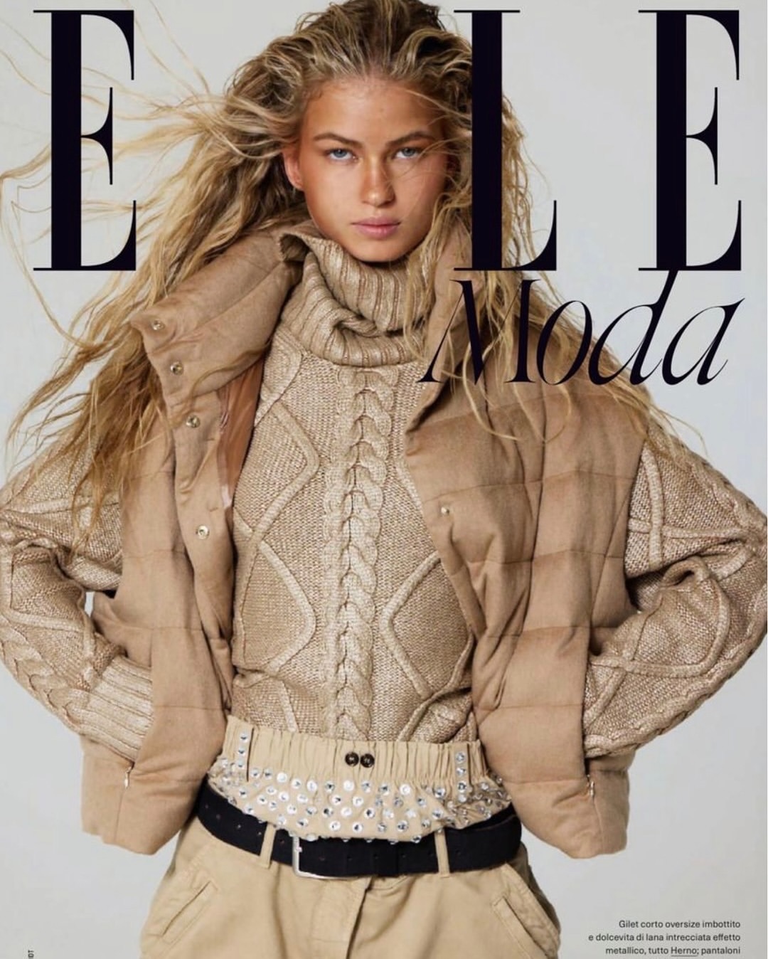 Close-up fashion editorial portrait of a woman with tousled blonde hair wearing a cable-knit turtleneck sweater and padded vest.