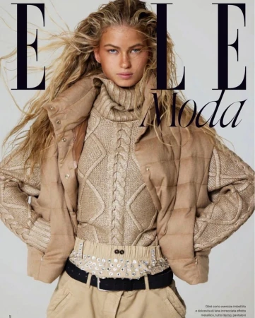 Close-up fashion editorial portrait of a woman with tousled blonde hair wearing a cable-knit turtleneck sweater and padded vest.