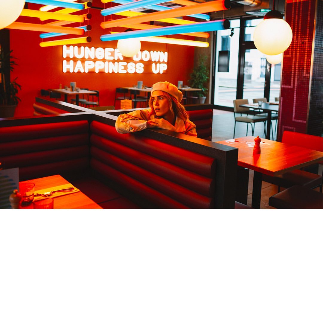 A woman in a restaurant booth gazes off with a curious expression. The restaurant's vibrant interior features a glowing neon sign.