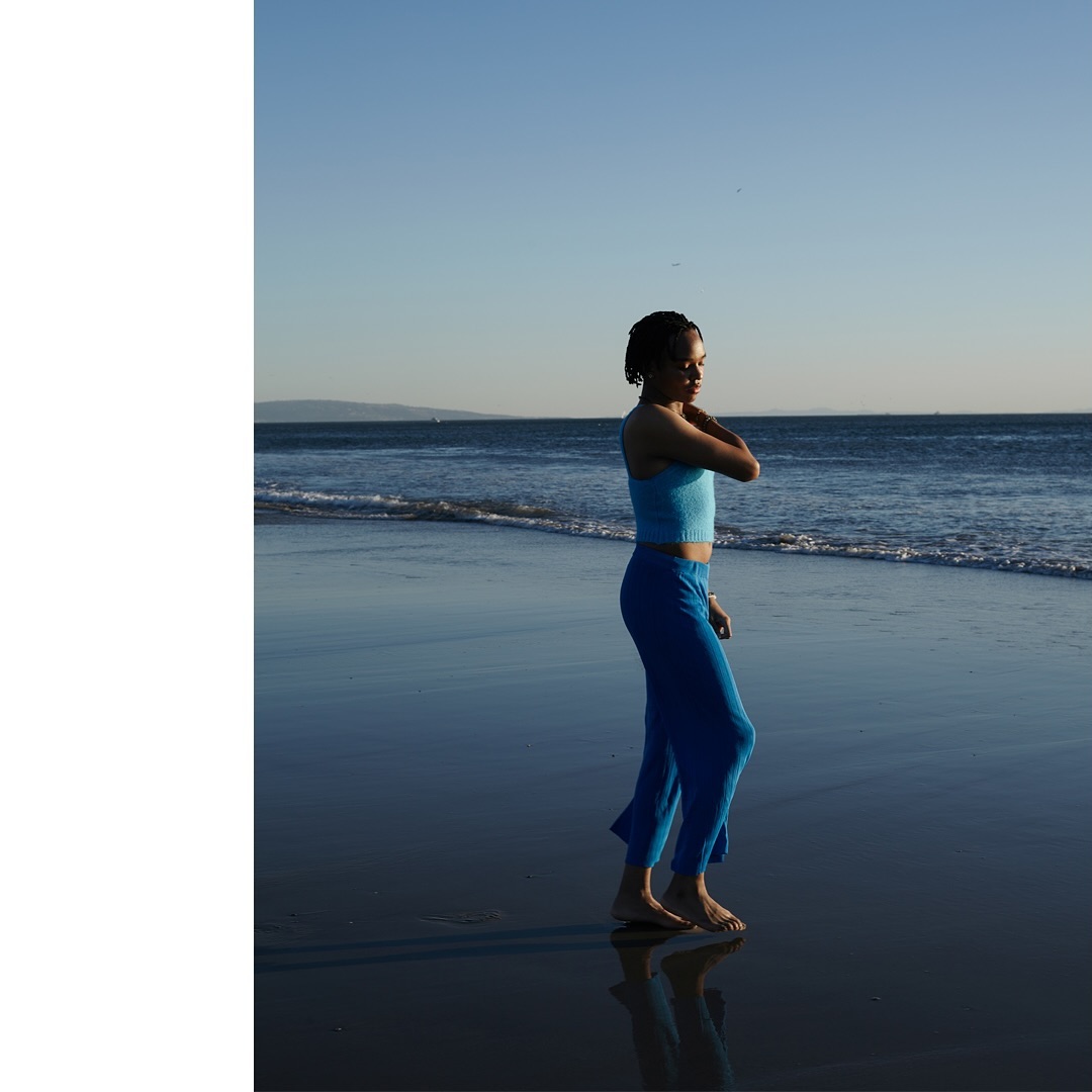 Adult female in blue athletic wear stands barefoot on a reflective beach at dusk with a blue sky and distant ocean waves.