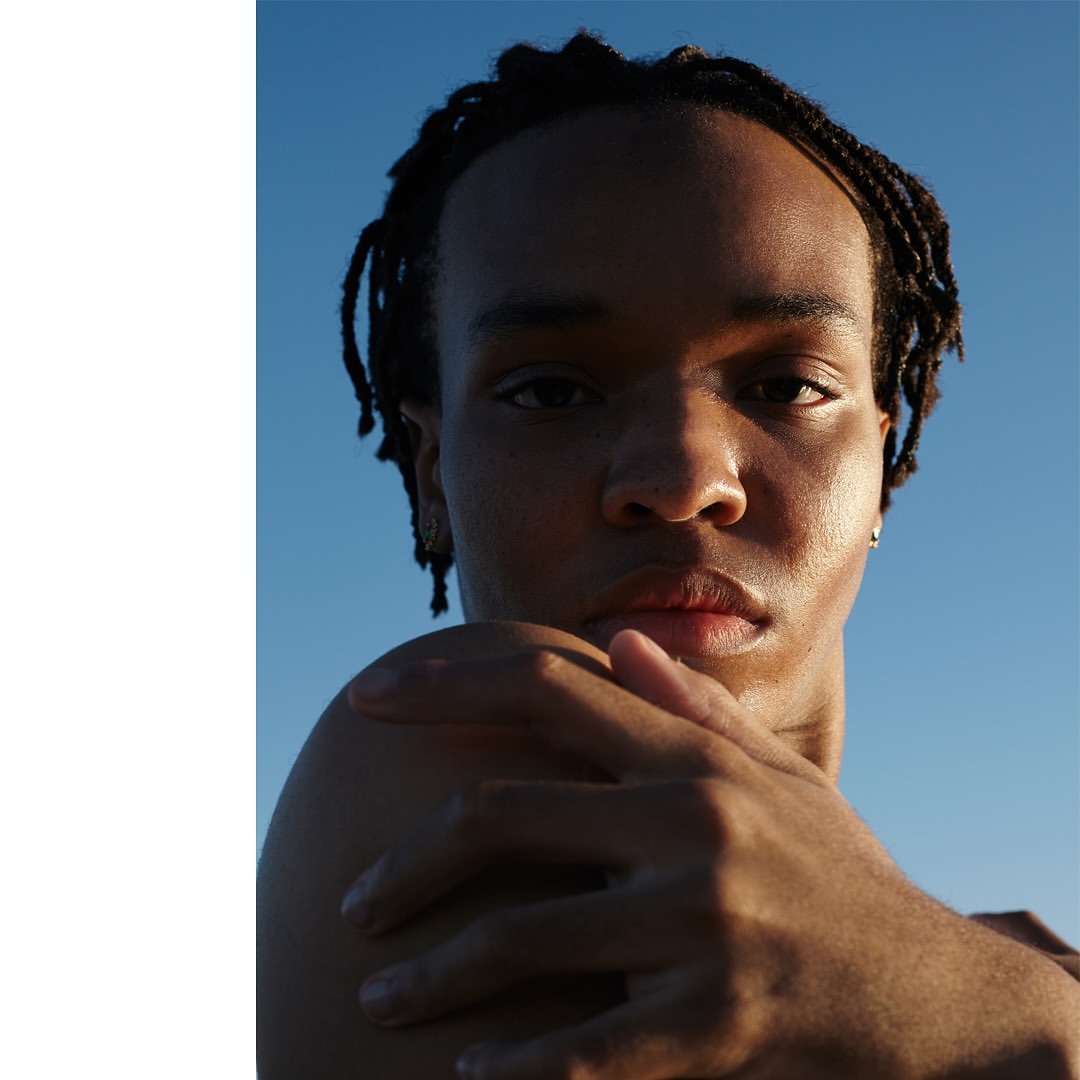 Close-up portrait of a bare-chested person with braided hair, arms crossed against a clear blue sky, bathed in soft, warm light.