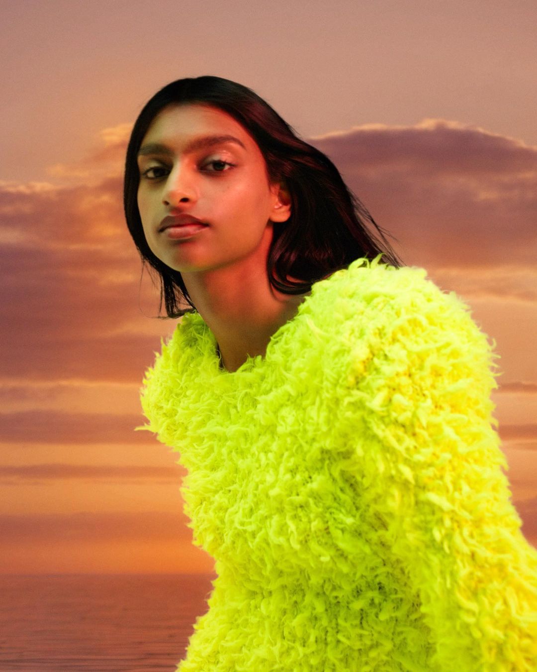 Woman with dark hair poses in a bright yellow, textured jacket against a sky with orange and pink clouds at sunset.