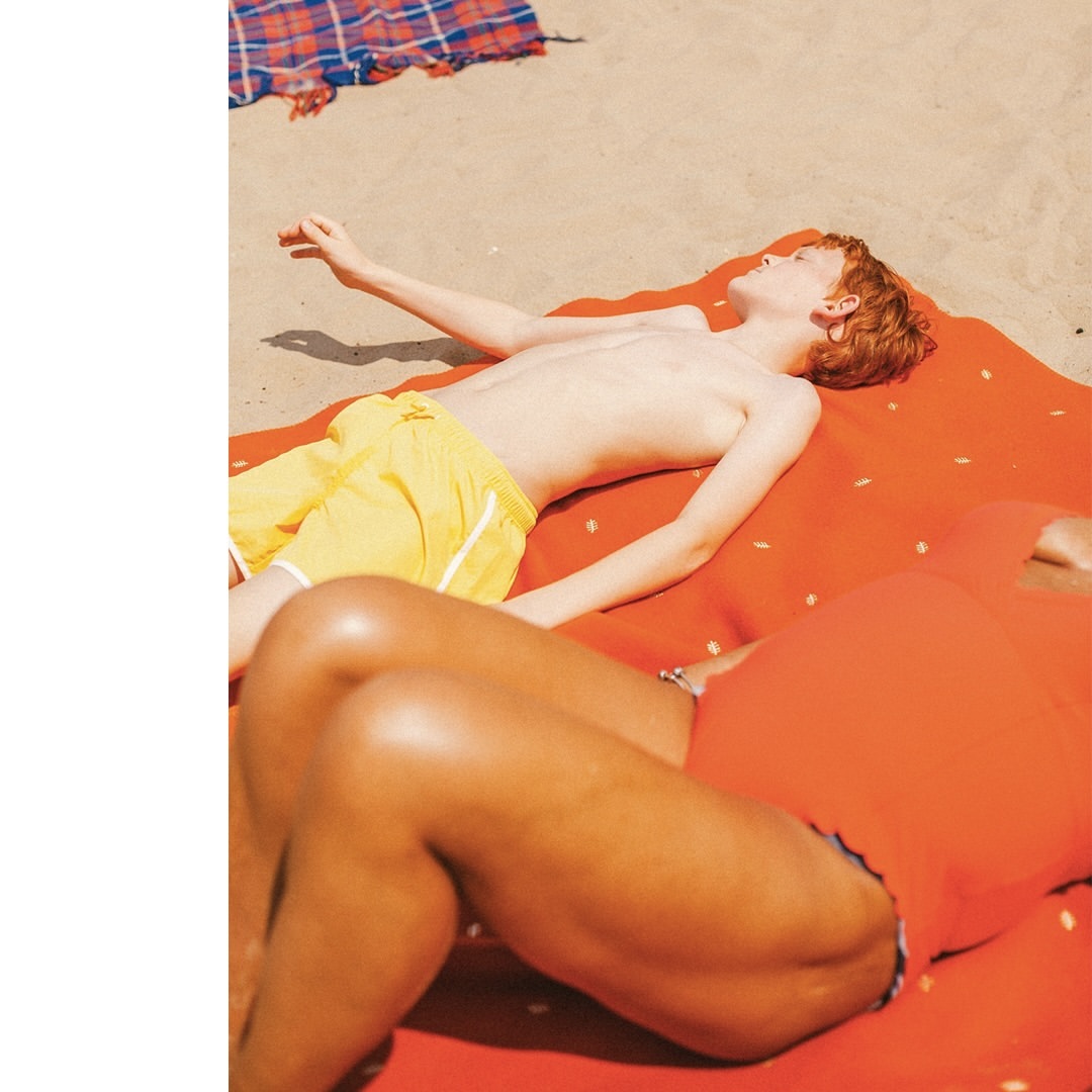 Two people sunbathing on an orange blanket on a sandy beach. One is a fair-skinned male with red hair and the other is a woman.