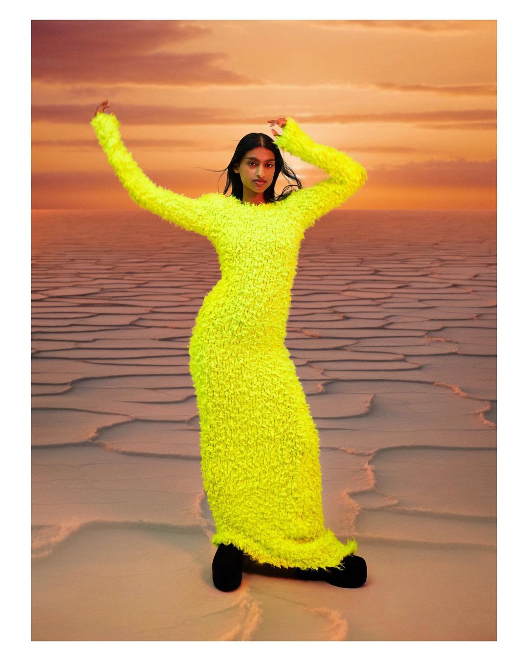 Fashionable woman in a long, bright yellow sweater dress poses in a desert landscape with a sunset in the background.