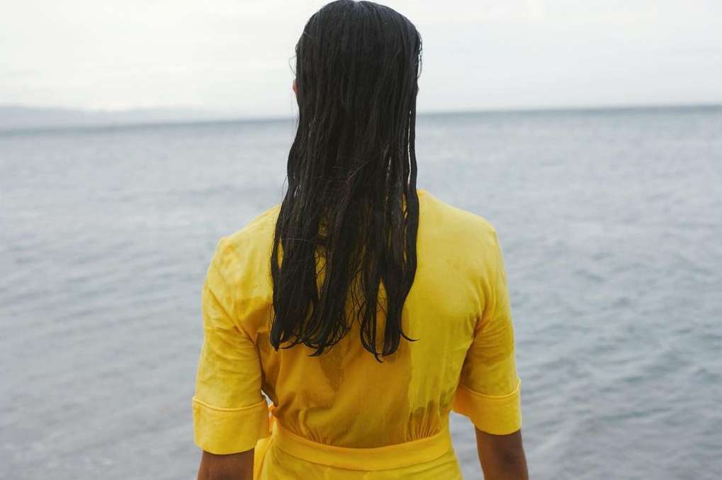 Back view of a woman with long dark hair wearing a yellow dress, standing near the ocean, looking out at the calm water and cloudy sky.