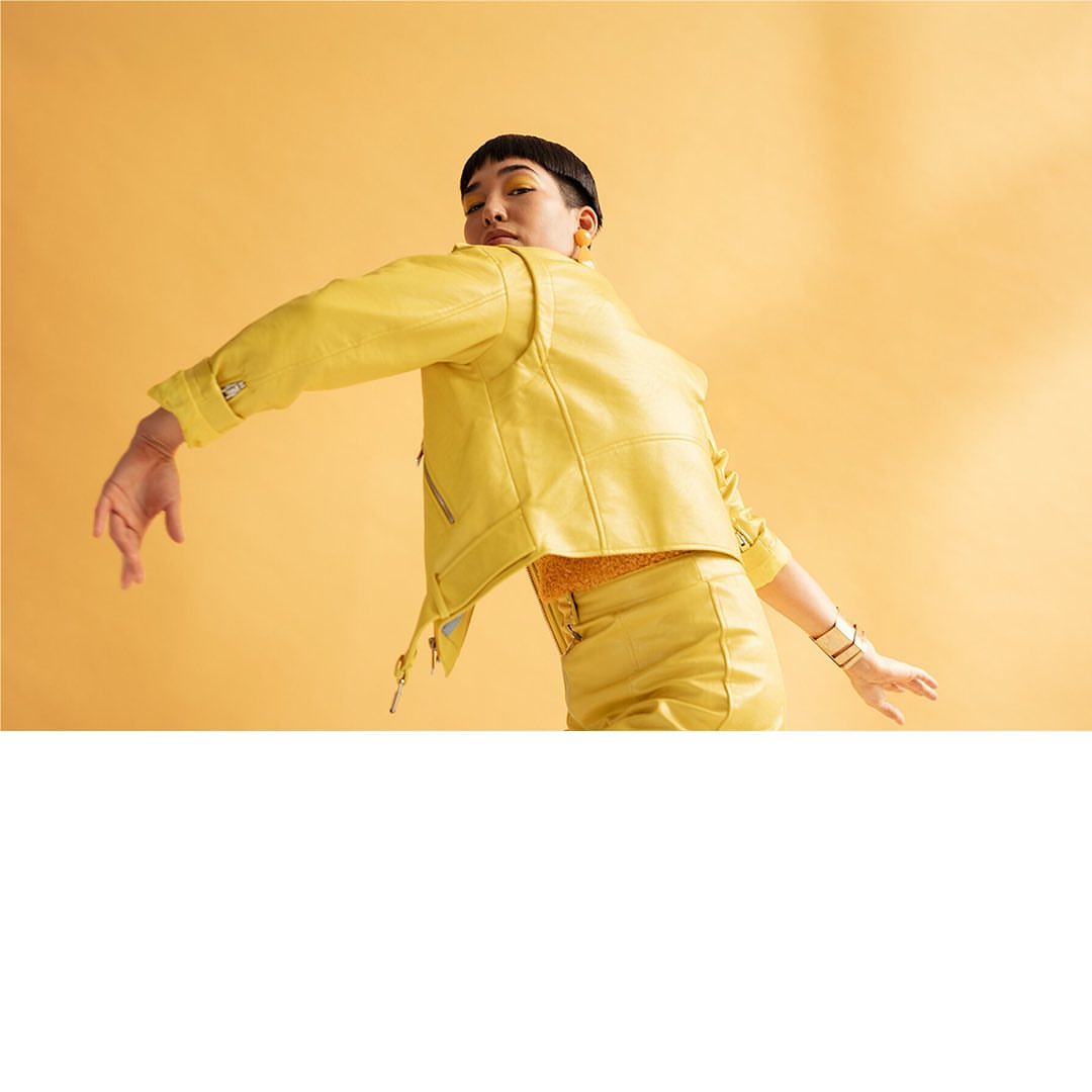 Eye-level shot of a person with a yellow jacket and skirt, posing with outstretched arms against a matching seamless backdrop.