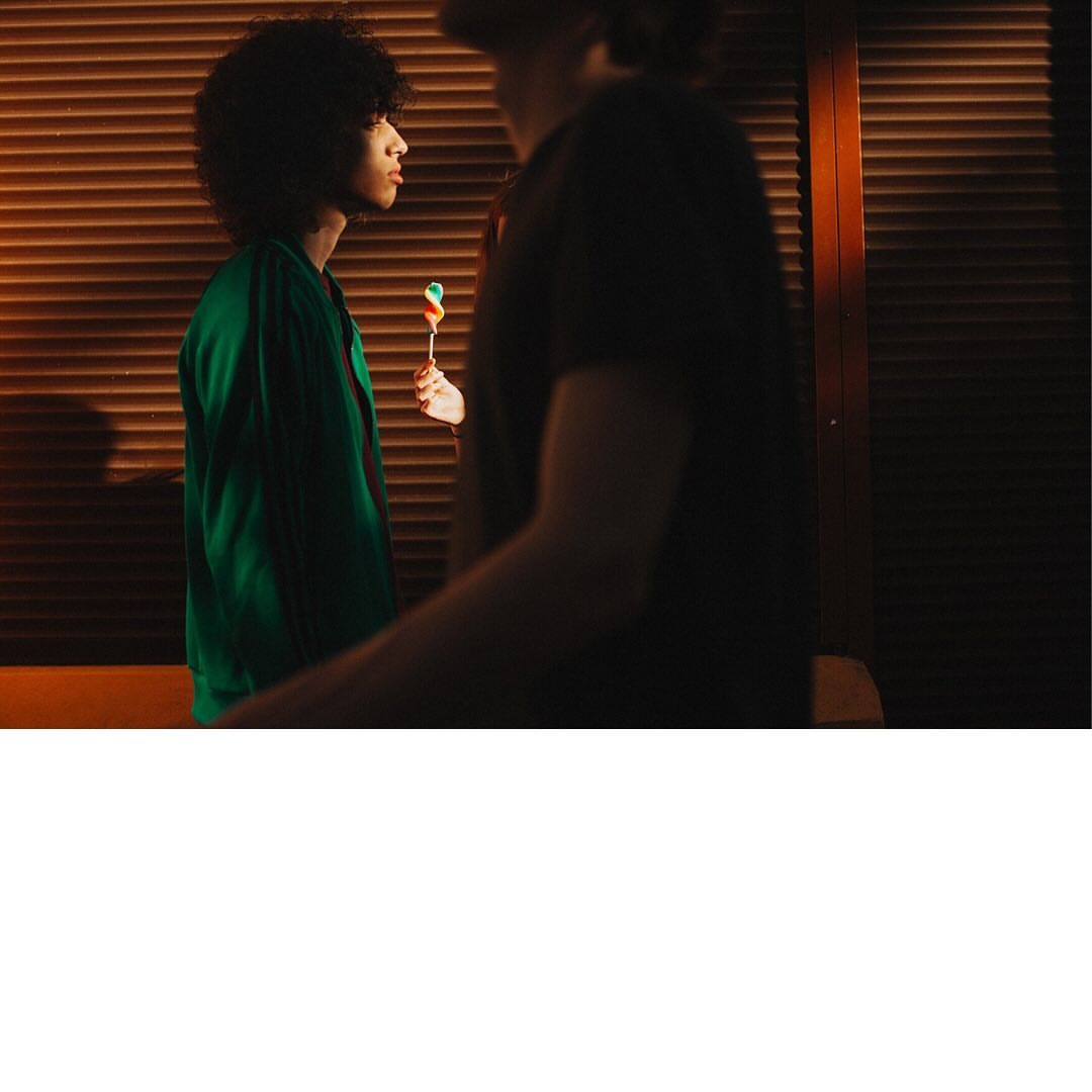 Two people indoors. One young adult with an afro holds a lollipop; the other, partially visible, stands next to wood blinds.