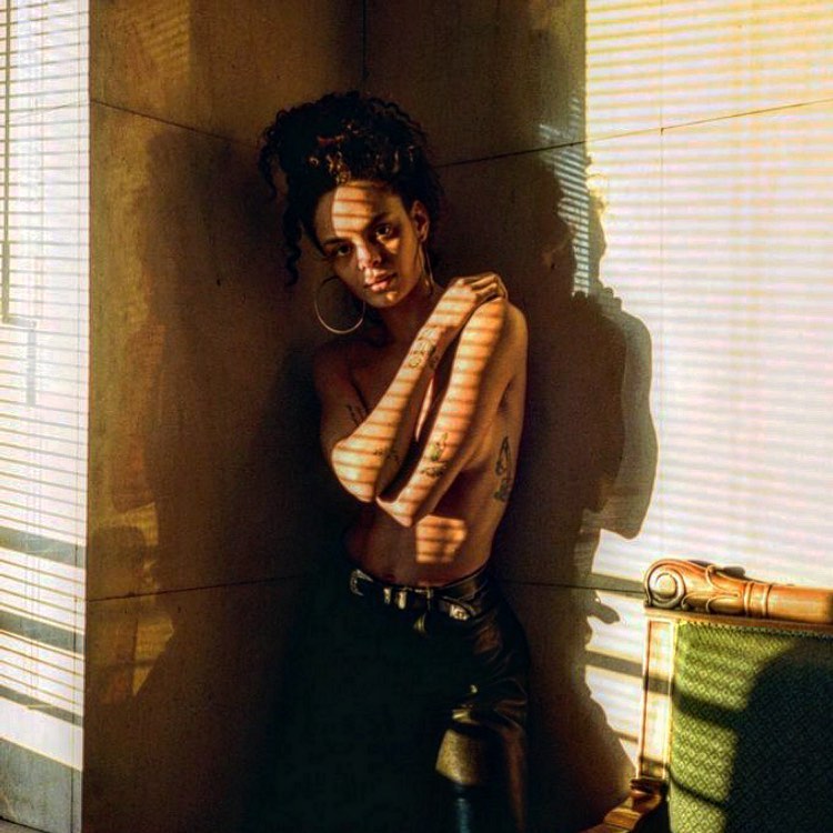 A young adult woman with curly dark hair poses in a corner with sunlight streaming through window blinds, casting striped shadows.