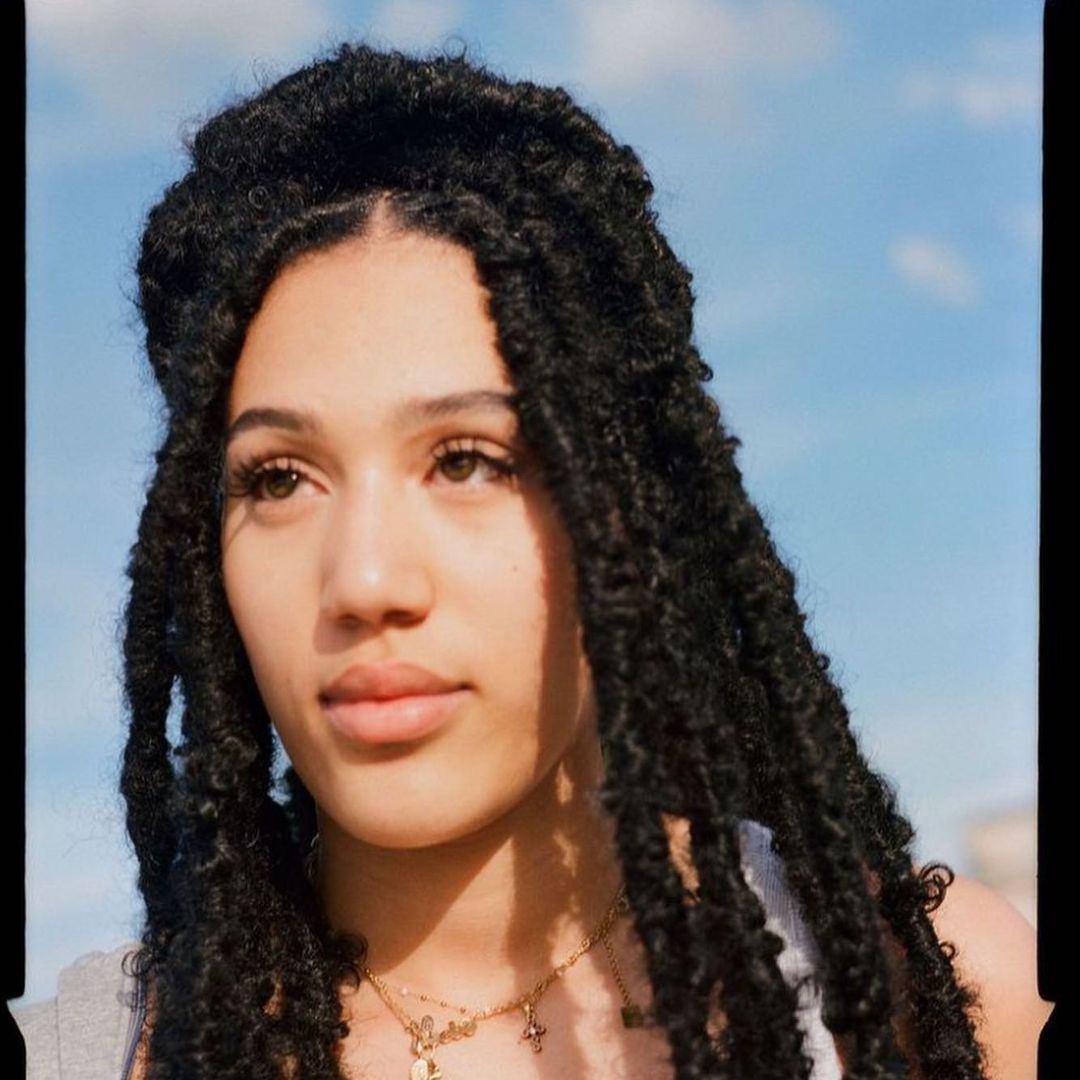 Portrait of a young woman with dark braided hair against a bright blue sky, wearing a grey top and gold necklaces.