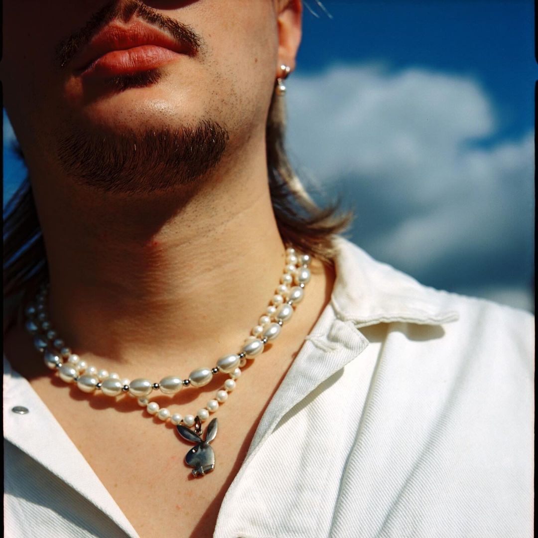 Close-up portrait of adult male with a pearl necklace against a bright blue sky with soft clouds, wearing a white button-down shirt.