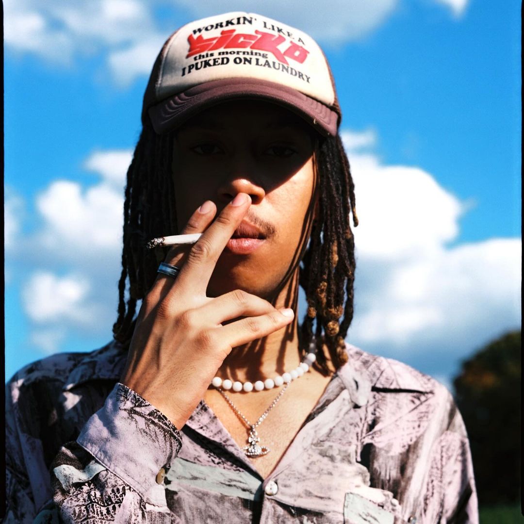 Young adult with dreadlocks and a baseball cap is smoking outdoors against a blue sky. Shirt has a light pattern and accessories.