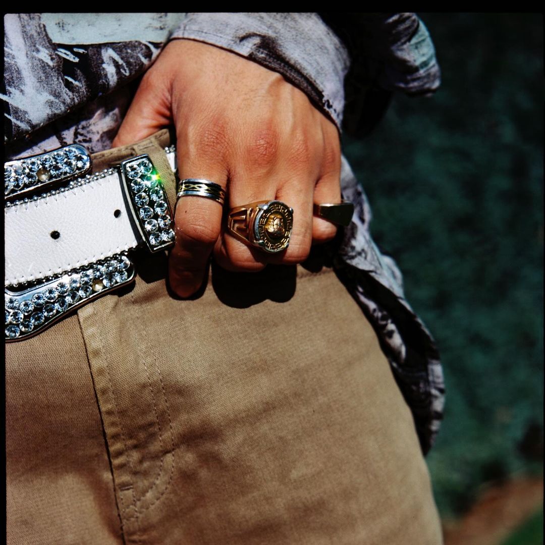 Close-up of a person's hand adorned with multiple rings, stylishly accessorized with a white, jewel-studded belt and a patterned shirt.