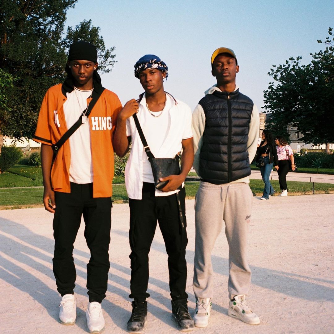 Three young adult Black men stand in a park wearing streetwear fashion with trees and other people in the background during daylight.